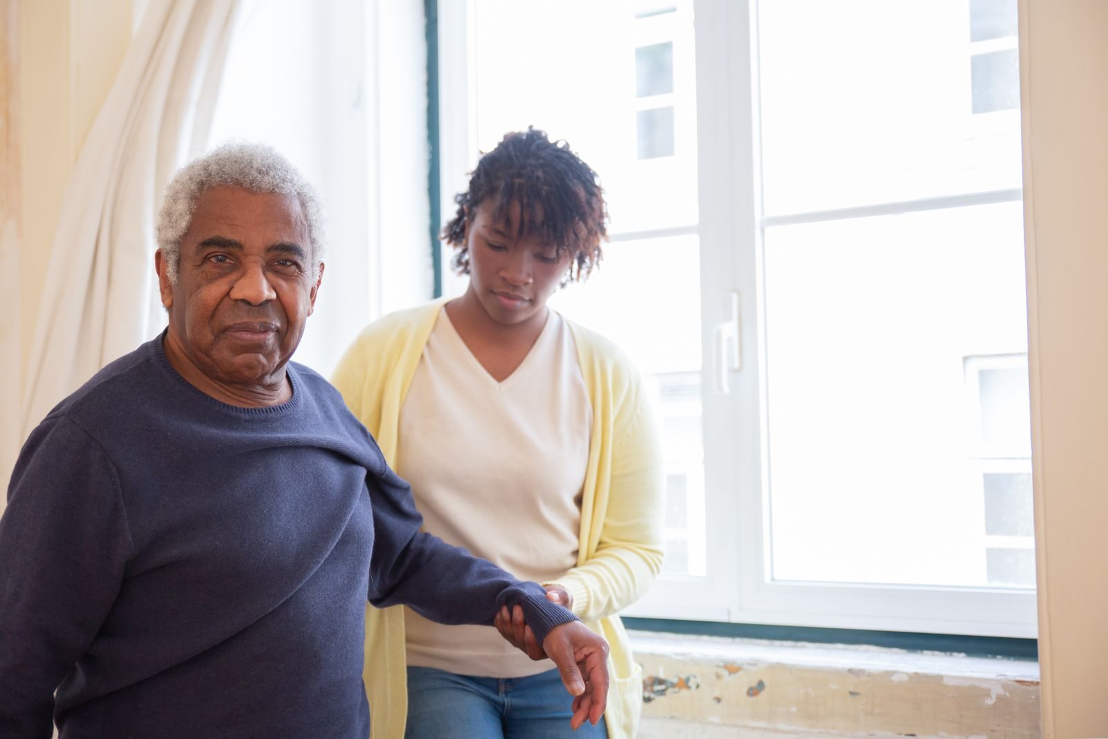 A home health aide walking with an older man by a bright window